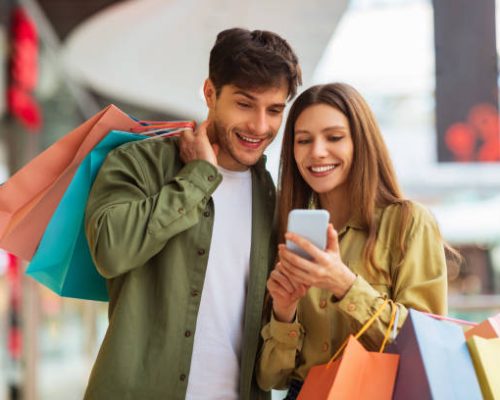 Happy Couple Shopping Using Mobile Phone Application Holding Colorful Shopper Bags And Buying Clothes In Modern Mall On Weekend. Customers Hold Smartphone Browsing Internet For Sales. Ecommerce